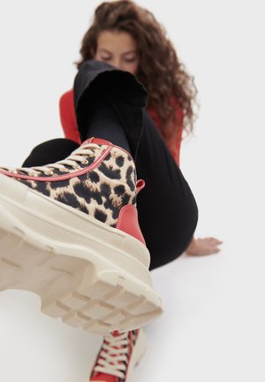 Nesreće s uzorkom leoparda, visoki tenisice s crvenim detaljima, teksturirani platneni gornji dio i debeli, rebrasti bijeli potplat. Oči i vezice u zlatnoj boji.