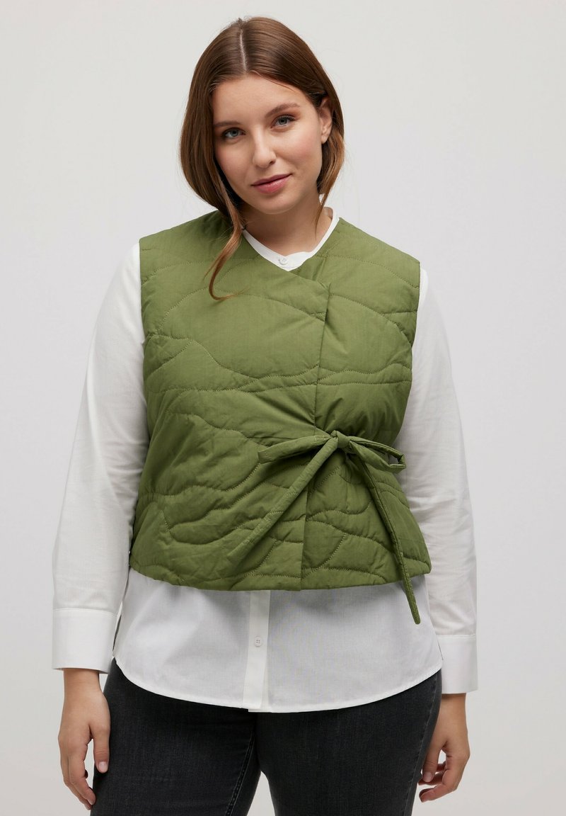Grüne, gesteppten Weste mit einem Bindeband in der Taille, die über einem weißen Langarmhemd getragen wird. Der Stoff hat eine glatte Textur und ein einfaches Design.