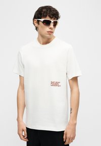 Νέος άνδρας που φοράει λευκό γραφικό t-shirt και λευκά αθλητικά γυαλιά ηλίου, στέκεται μπροστά σε απλό ανοιχτό φόντο, κοιτώντας πλάγια.