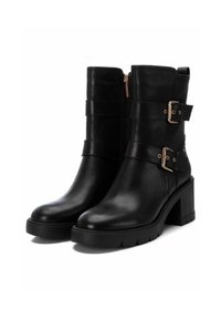 Botas de tobillo de cuero negro con un tacón grueso, que presentan dos correas anchas y hebillas de metal. Textura suave y cierre de cremallera en el lateral.