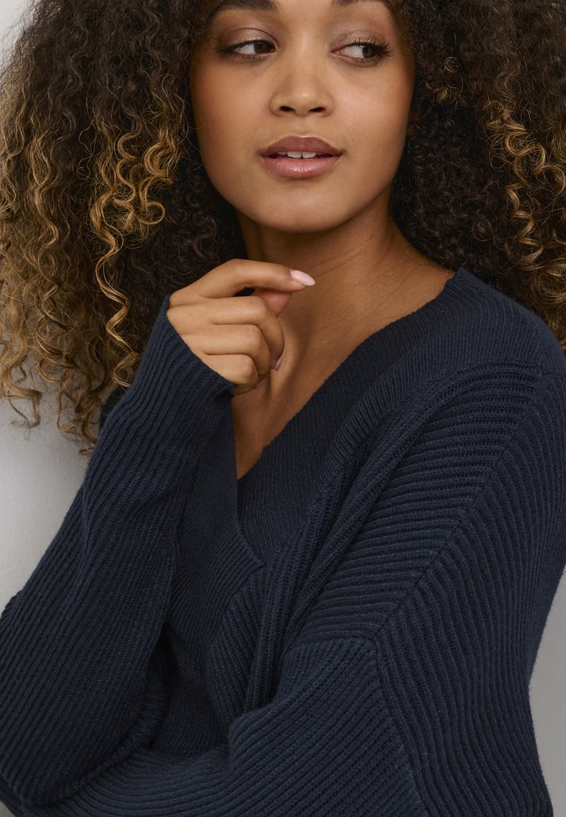 Käharate juustega naine tumel sinises soonikuga kampsunis, kes vaatab küljele, käsi lõua lähedal, tavalise tausta vastu.