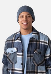 Chemise en laine à carreaux bleus et gris avec deux poches poitrine et des boutons noirs, portée sur un t-shirt bleu clair et un bonnet en laine gris.