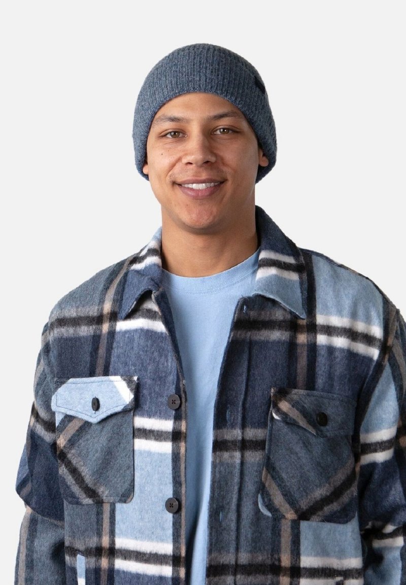 Chemise en laine à carreaux bleus et gris avec deux poches poitrine et des boutons noirs, portée sur un t-shirt bleu clair et un bonnet en laine gris.