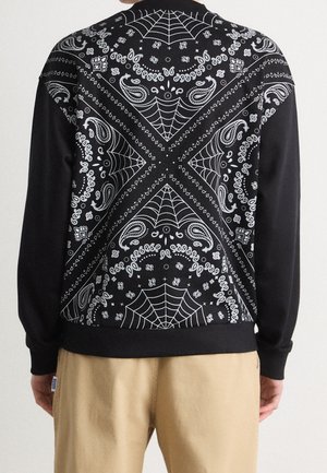 Schwarzes langärmliges Hemd mit weißen Paisley- und Spinnennetz-Bandana-Mustern, getragen von einer Person in beigen Hosen, von hinten gezeigt.
