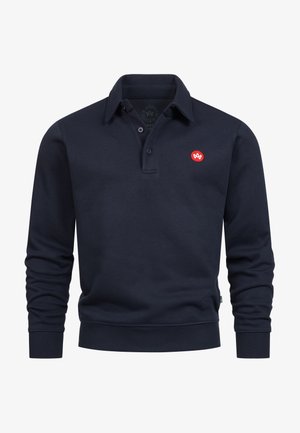 Marineblå bomulls-pikéskjorte med lange ermer, ribbestrikkede mansjetter og krage. Har en liten rød sirkel-logo på brystet. Enkelt design.