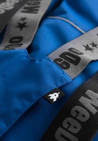 Blått tyg med slät textur, som har en svart logotypetikett och grå remmar med djärva svarta textmönster. Närbild på söm och design.