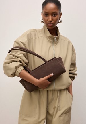 Femme en combinaison beige à fermeture éclair tenant un sac à main rectangulaire marron foncé avec deux poignées sur un fond uni.