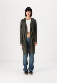 Lange olijfgroene cardigan met een open voorzijde, zachte textuur en twee zijvakjes, gedragen over een witte crop top en blauwe jeans. Zwarte sneakers.