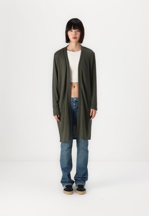Lång olivgrön kardigan med öppen framsida, mjuk textur och två sidofickor, som bärs över en vit crop top och blå jeans. Svarta sneakers.
