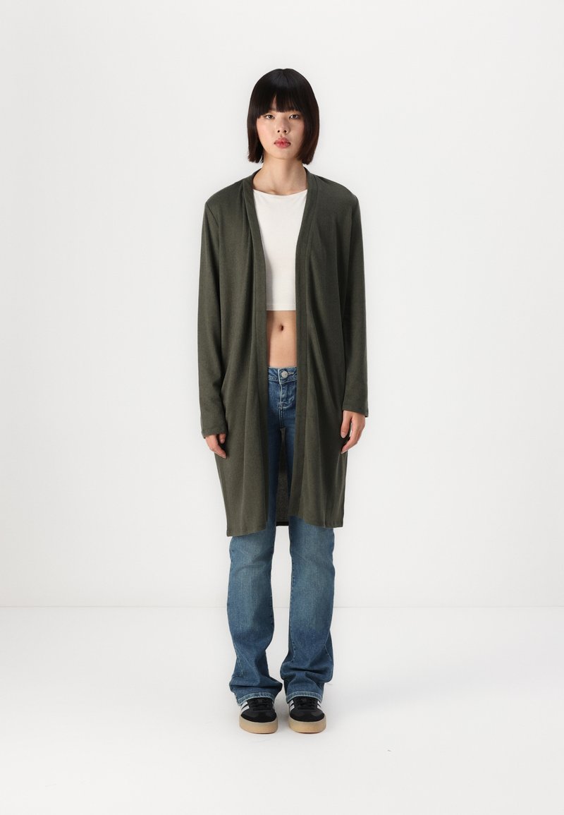 Lange olijfgroene cardigan met een open voorzijde, zachte textuur en twee zijvakjes, gedragen over een witte crop top en blauwe jeans. Zwarte sneakers.