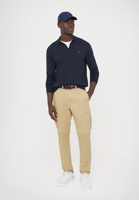 Temno modra polo majica z dolgimi rokavi, kaki hlače z žepi na straneh, beli supergi in temno moder šepek z logotipom.