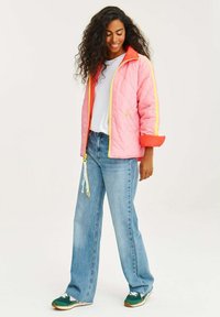 Prošívaná růžová bunda s oranžovým límcem a žlutými pruhy, zkombinovaná s světle modrými širokými džíny a zelenými teniskami s barevnými akcenty.