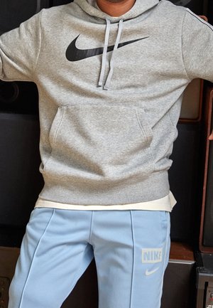 Grå hoodie med et sort Nike-logo, der har en frontlomme og snøre. Sammen med lyseblå bukser, der viser "NIKE" logoet.