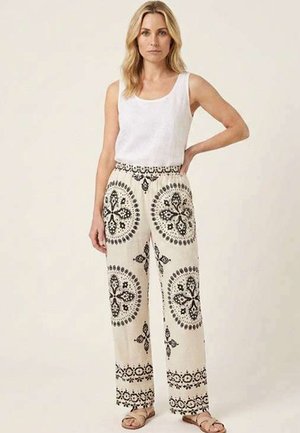 Donna con capelli biondi che indossa una canotta bianca senza maniche, pantaloni crema a gamba larga con motivi circolari neri e sandali beige, in piedi con una mano sui fianchi.
