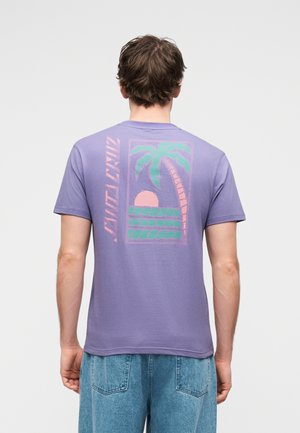 Férfi, aki levendula színű pólót visel, amelyen pálmafás, naplementés és vízszintes csíkos minta található a hátán, világoskék farmerral párosítva.