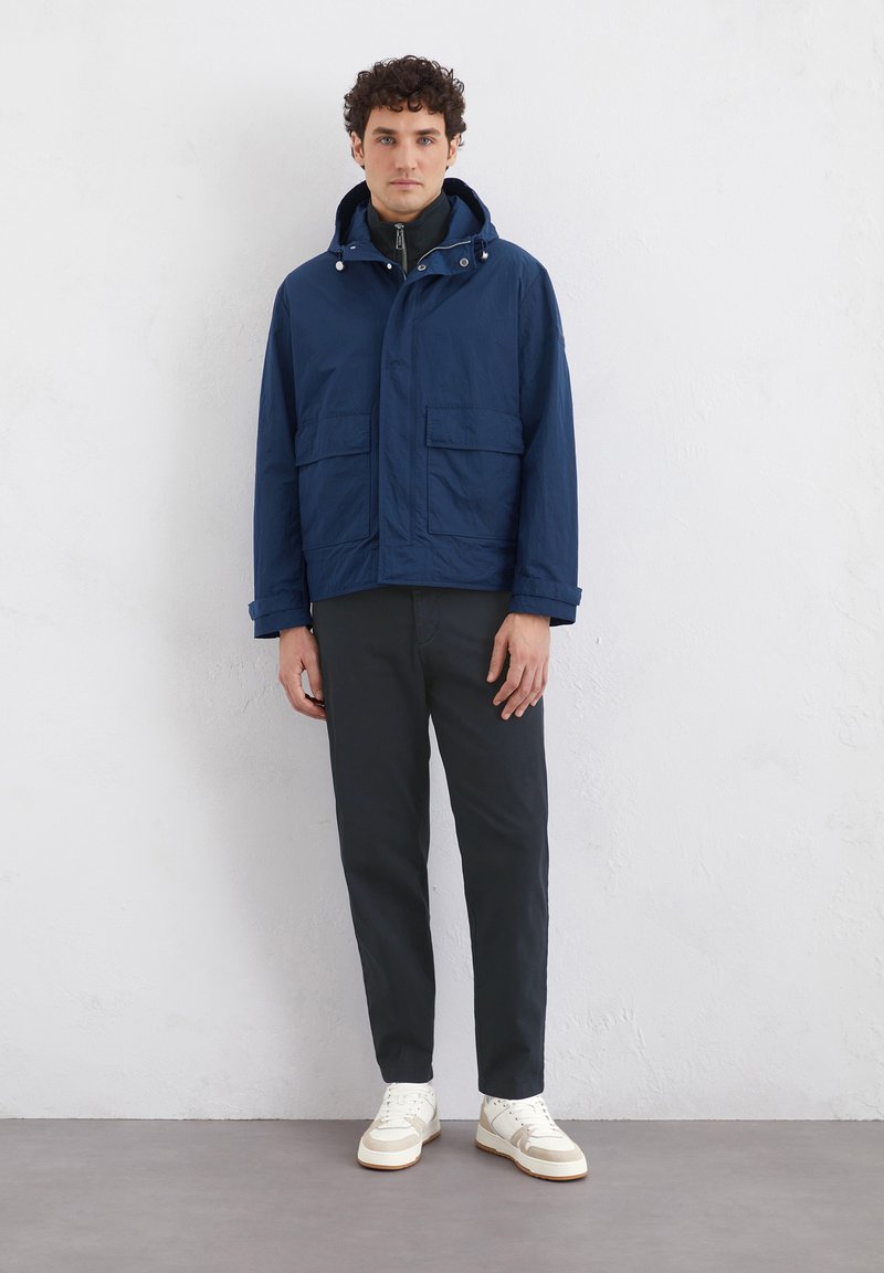 Marineblauwe waterdichte jas met twee voorzakken, verstelbare capuchon en ritsen. Gecombineerd met donkere broeken en lichte sneakers.