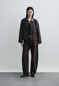 Bruin patroonjack met grote zakken, contrasterende kraag, gecombineerd met donkere, loszittende broek en zwarte schoenen, staand op een grijze vloer.