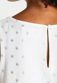 Haut en tissu blanc texturé avec des pois argentés. Le dos présente une ouverture en forme de trou de serrure et une fermeture à bouton. Matière lisse et légère.