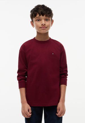 Garçon aux cheveux courts et foncés portant une chemise bordeaux à manches longues et un pantalon foncé, debout devant un fond blanc uni.