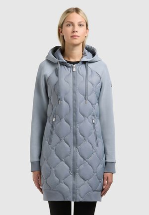 Vrouw draagt een lange, lichtblauwe doorgestikte jas met capuchon en ritssluitingen aan de zakken, staande tegen een effen achtergrond.