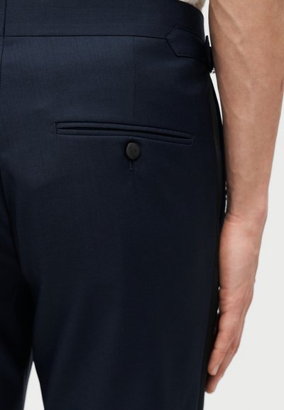 Pantaloni croiți bleumarin închis, cu un singur buzunar în spate fixat cu un nasture negru, purtați de o persoană cu brațul relaxat pe laterală.