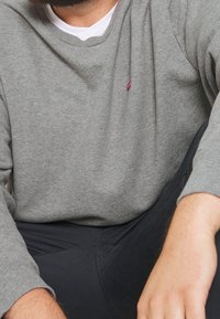 Homme portant une chemise grise à manches longues sur un sous-vêtement blanc et un pantalon foncé, assis avec un bras reposant sur son genou.