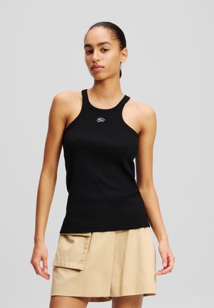 Black ribbed tank top with a round neckline and narrow straps, featuring a small logo in the center. Paired with a beige skirt.