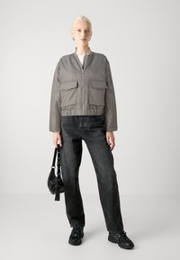 Grå bomberjakke med frontlommer, kombinert med sorte rettsnittjeans og svarte joggesko. Modell som holder en liten svart håndveske.