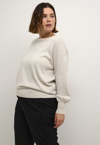 Pull beige clair à la coupe décontractée, texture douce, manches larges et poignets élastiques. Assorti à un pantalon noir.