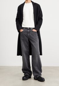 Manteau noir, t-shirt blanc et jeans gris ample. La tenue présente une texture lisse et une silhouette classique avec des chaussures noires robustes.
