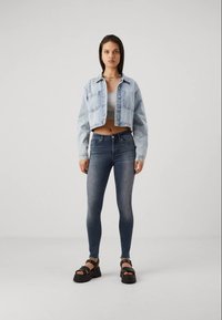 Lys blå denimjakke med avkuttede ermer over en grå topp, med mørk blå skinny jeans og kraftige svarte sandaler, mot en ensfarget bakgrunn.