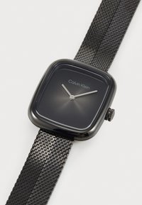 Montre carrée noire avec une finition brillante, bracelet en mailles et aiguilles argentées minimalistes. Présente un cadran noir et le nom de la marque affiché.