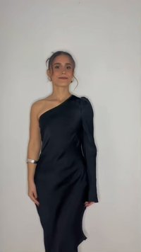 Femme aux cheveux foncés portant une robe noire une épaule, debout devant un fond clair uni, regardant devant elle avec un léger sourire.
