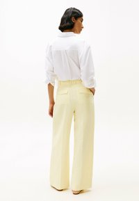 Camisa blanca de botones con mangas arremangadas, combinada con pantalones de tiro alto y perneras anchas de color amarillo pálido que tienen bolsillos traseros y una textura suave.