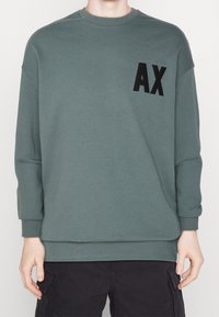 Sweatshirt i dämpad grön med rundad halsringning, långa ärmar, ribbade muddarna och en distinkt svart "AX"-logotyp på framsidan.