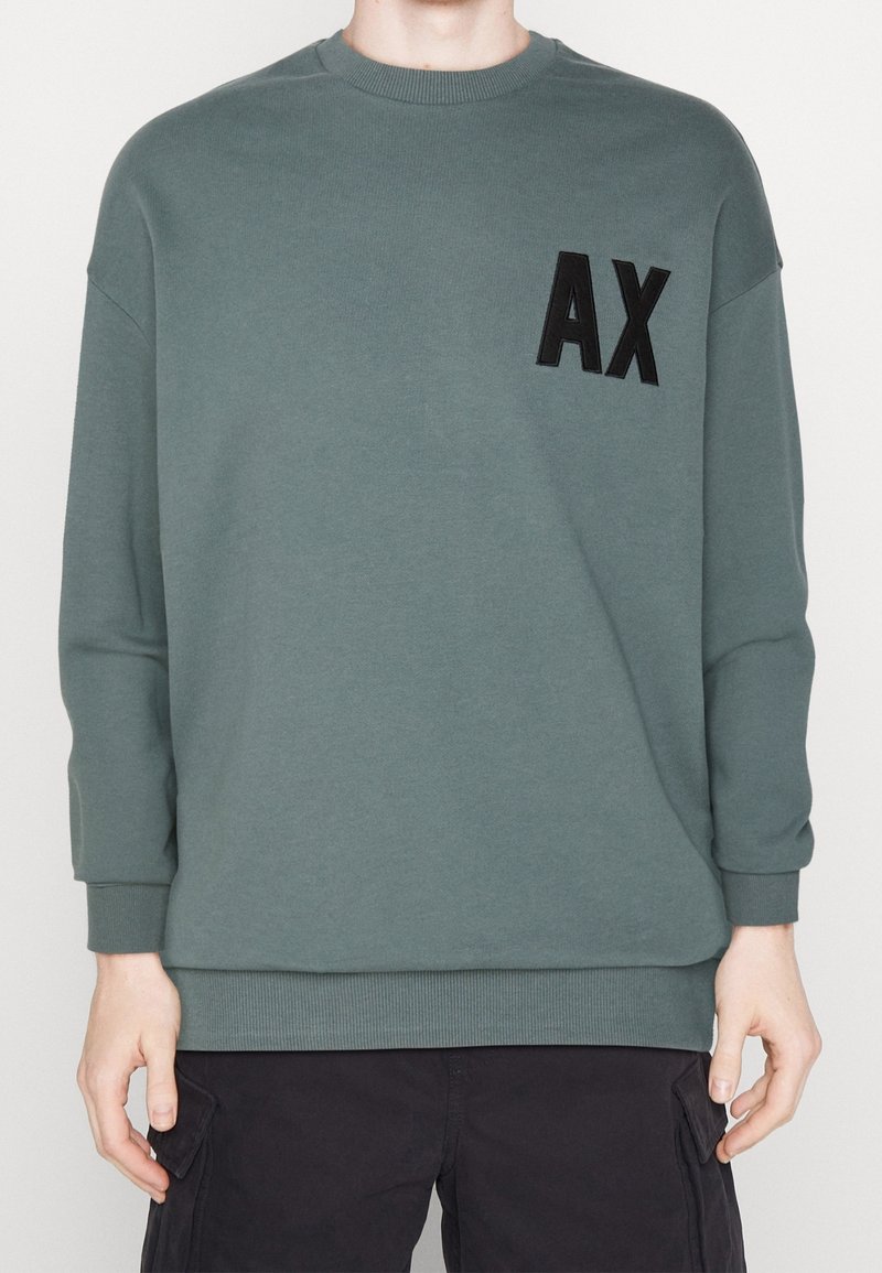 Sweatshirt i dämpad grön med rundad halsringning, långa ärmar, ribbade muddarna och en distinkt svart "AX"-logotyp på framsidan.
