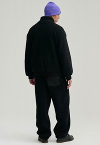 Czarna polarowa kurtka z wysokim kołnierzem, zestawiona z dopasowanymi czarnymi polarowymi spodniami. Uzupełniona fioletową dzianą czapką i czarnym obuwiem.