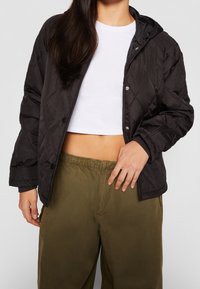 Schwarze gesteppte Jacke mit Kapuze, Druckknöpfen und gerippten Bündchen, getragen über einem weißen Crop-Top und olivgrünen Hosen mit elastischem Bund.