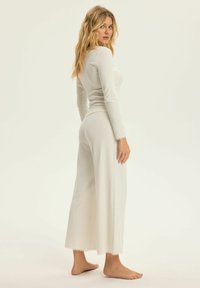 Kobieta z długimi blond włosami, ubrana w białą, prążkowaną bluzkę z długim rękawem oraz spodnie z szerokimi nogawkami, stoi boso na tle jednolitym.