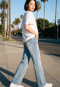 Giovane donna con t-shirt bianca e jeans azzurri che cammina su una strada soleggiata fiancheggiata da palme, guardando sopra la spalla verso la fotocamera.