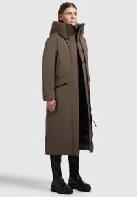 Kvinde iført lang brun polstret frakke med høj krave, stående med hænderne ned langs siden, parret med sorte kraftige støvler mod ensfarvet baggrund.