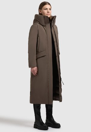 Ženska v dolgem rjavem oblazinjenem plašču z visokim ovratnikom, stoji z rokama ob straneh, obuta v črne robustne škornje, na enobarvni podlagi.