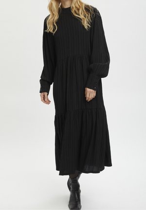 Femme portant une longue robe noire plissée avec des poignets boutonnés et des bottines noires, debout devant un fond uni.