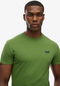 Grøn kortærmet t-shirt lavet af bomuld, med rund hals, flade sømme og et lille blåt logo på brystet.