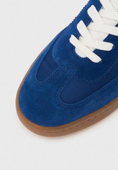 Kék velúr és hálós sportcipő fehér fűzőkkel, lekerekített orral és barna gumitalppal. Díszítő varrásokkal és kényelmes illeszkedéssel rendelkezik.