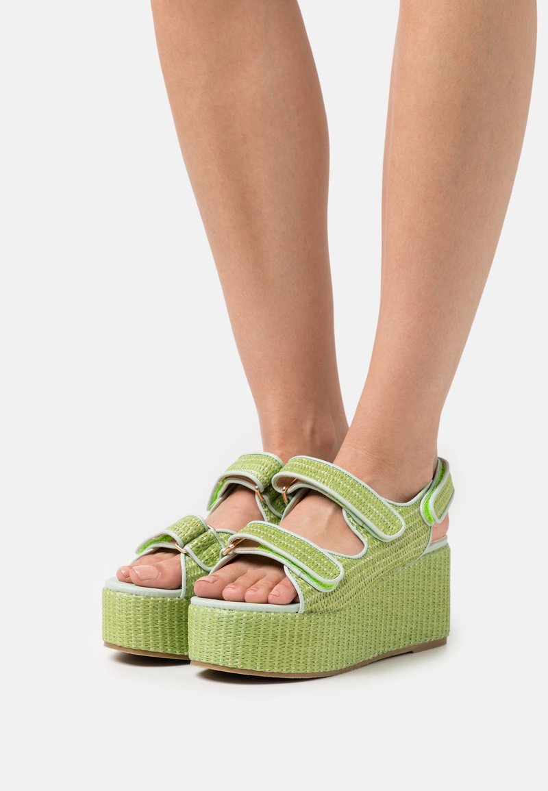 Pieds chaussés de sandales compensées vert clair avec deux brides réglables sur les orteils et une bride à la cheville, toutes avec une texture tissée.