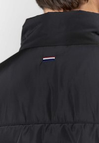 Baksidebilde av en person som har på seg en svart jakke med høy krage og en liten rød, hvit og blå stripete stoffetikett øverst på ryggen.