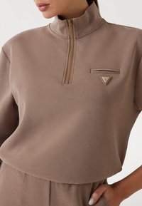 Lysebrun pullover med høj krave og halvdels lynlås, prydet med en lille lynlåslomme på brystet og en trekantet logoakkent på stoffet.