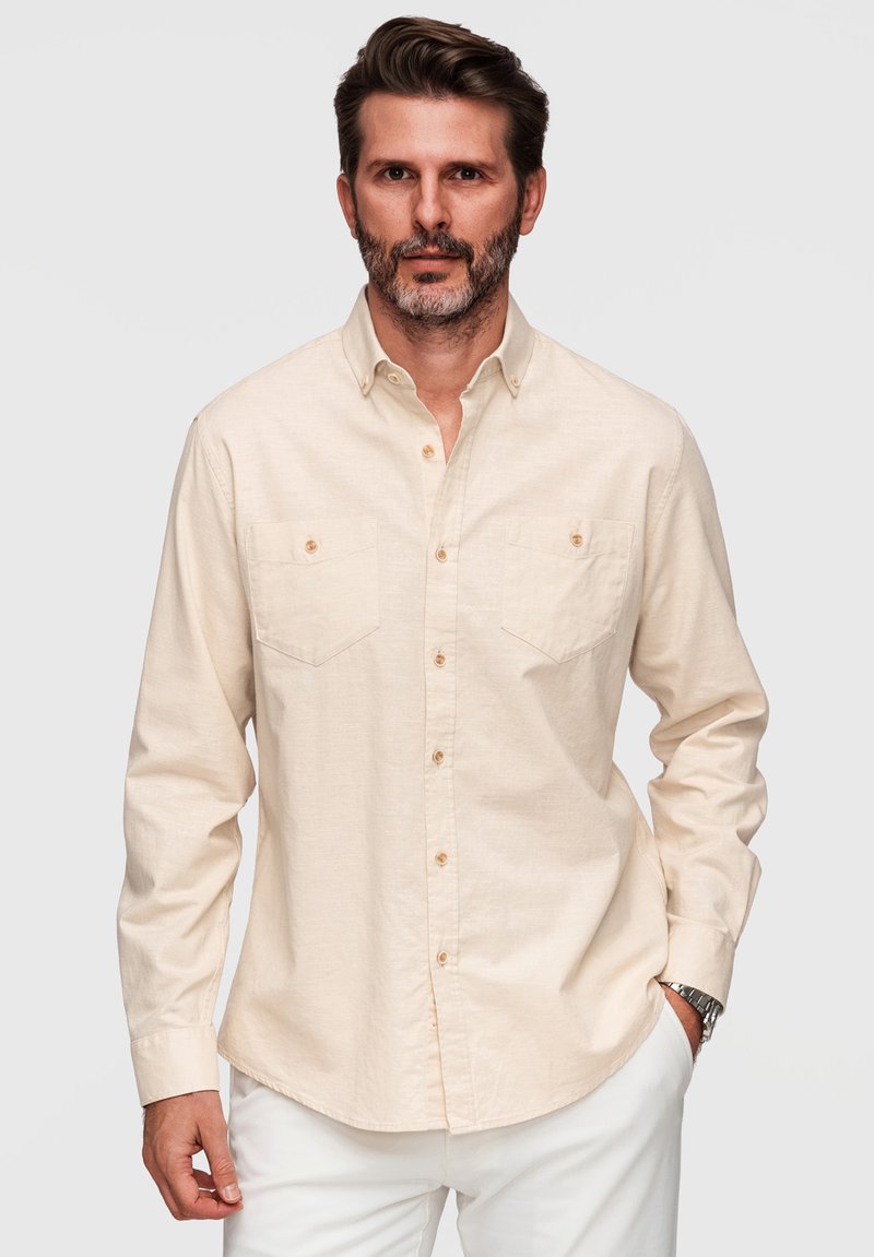 Homme aux cheveux bruns courts et à la barbe, portant une chemise beige à boutons avec des poches et un pantalon blanc, debout devant un fond uni.