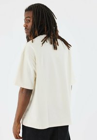 Camiseta de color crema, de corte holgado y con mangas cortas, de textura suave, combinada con pantalones negros sueltos, mostrando un ajuste relajado desde la parte de atrás.